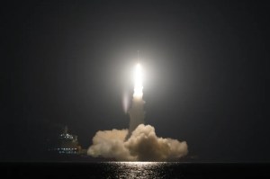 Nighttime sea launch of the Ceres-1 rocket, with bright exhaust illuminating the surrounding smoke and reflecting on the ocean surface. A mobile sea platform is visible in the background.