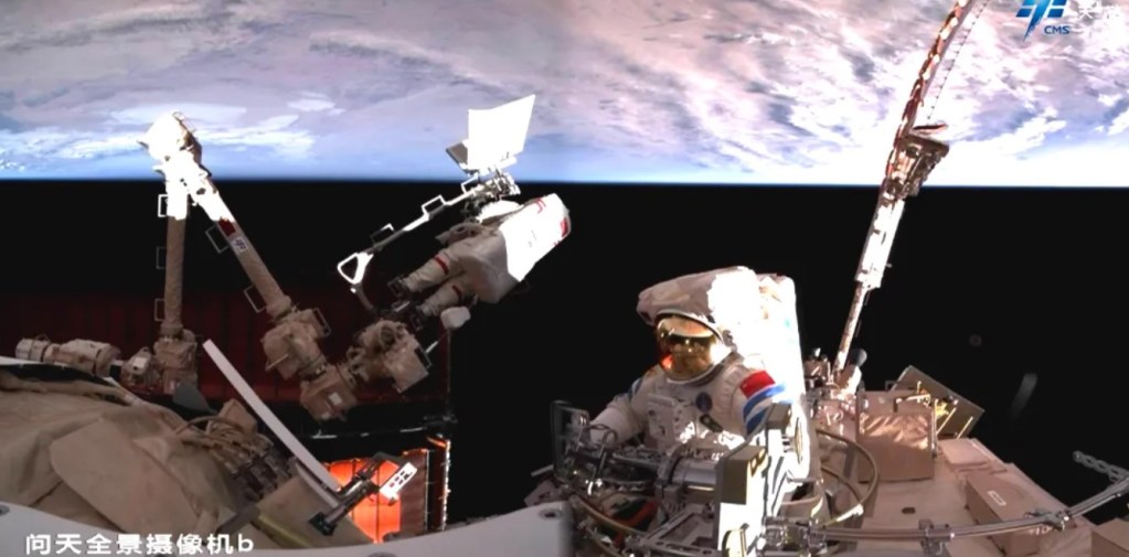 Two Chinese astronauts conduct extravehicular activity (EVA) outside the Tiangong space station, wearing Feitian spacesuits. One astronaut is attached to the station's robotic arm, working on external equipment, while the second astronaut is positioned near handrails with Earth’s curvature visible in the background. The image is captured by a panoramic camera on the Wentian module.