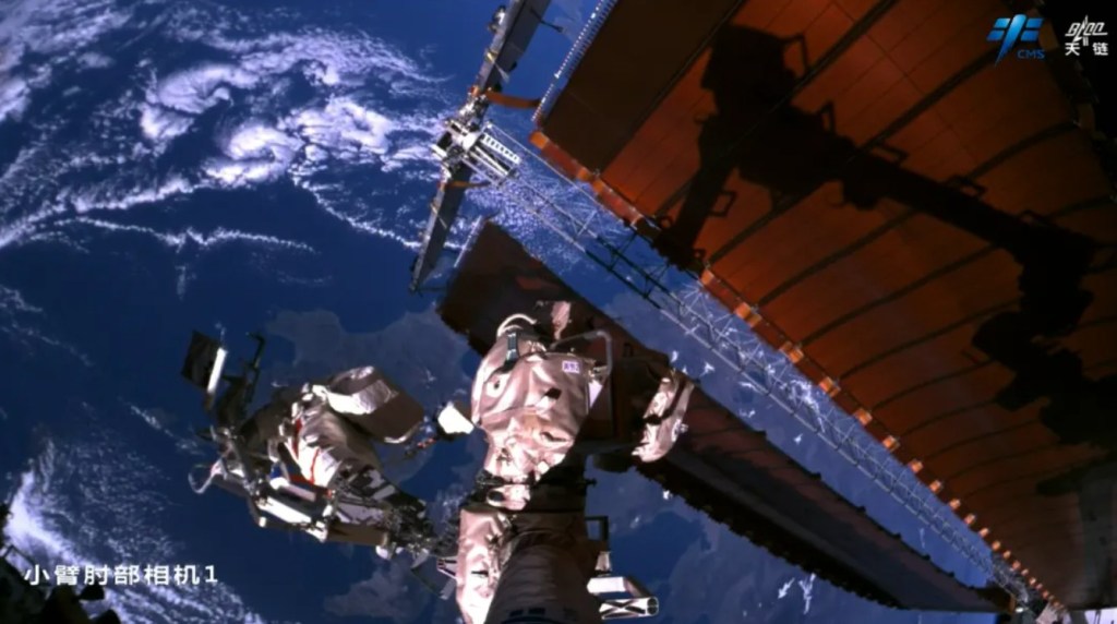 Two Chinese astronauts conducting a spacewalk outside the Tiangong space station, with Earth and clouds visible in the background. One astronaut is positioned at the end of Tiangong’s robotic arm, captured by an external camera mounted on the arm. Large solar arrays of the station are also visible.