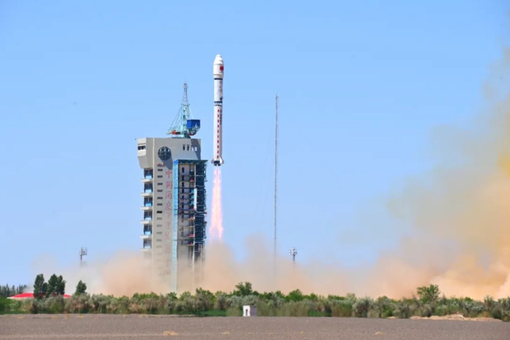 A white Long March 2D rocket lifts off from the Jiuquan Satellite Launch Center. The rocket ascends into a clear blue sky with exhaust plumes trailing below, as the launch tower and desert landscape appear in the foreground.