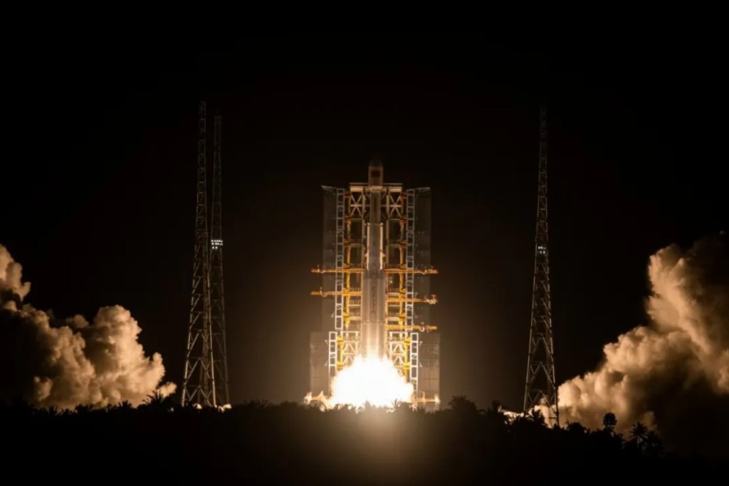 A Long March 7A rocket lifts off at night from China's Wenchang Satellite Launch Center, illuminating the launch pad with bright exhaust flames and casting shadows on surrounding support structures and trees.