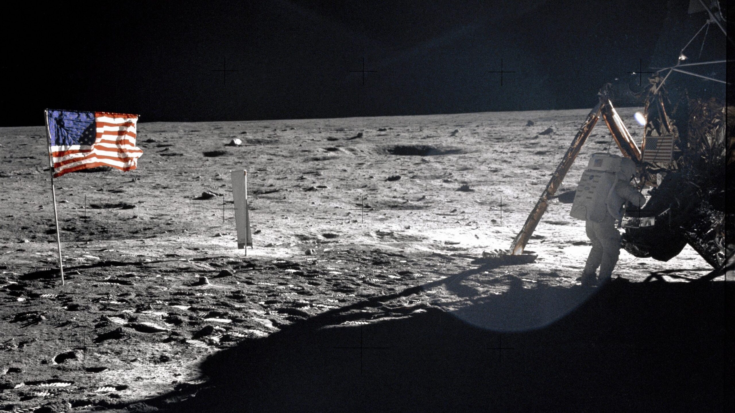 an astronaut in a white spacesuit descends from a gold-foil-wrapped spacecraft onto the gray lunar surface, with an american flag planted in the dirt nearby
