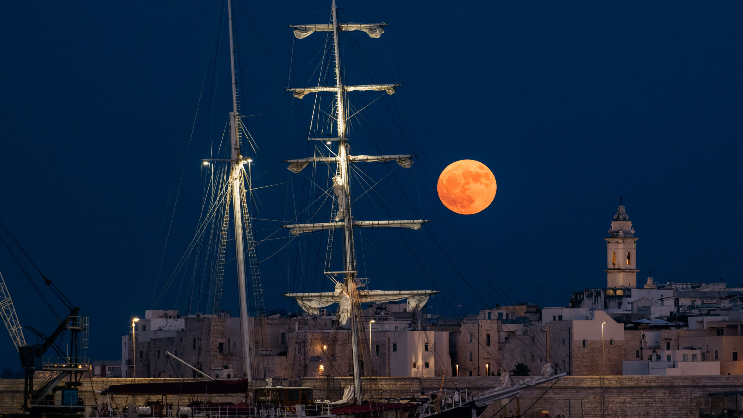 a large full moon with a pinkish orange hue sits low in the sky behind a large sailing boat.