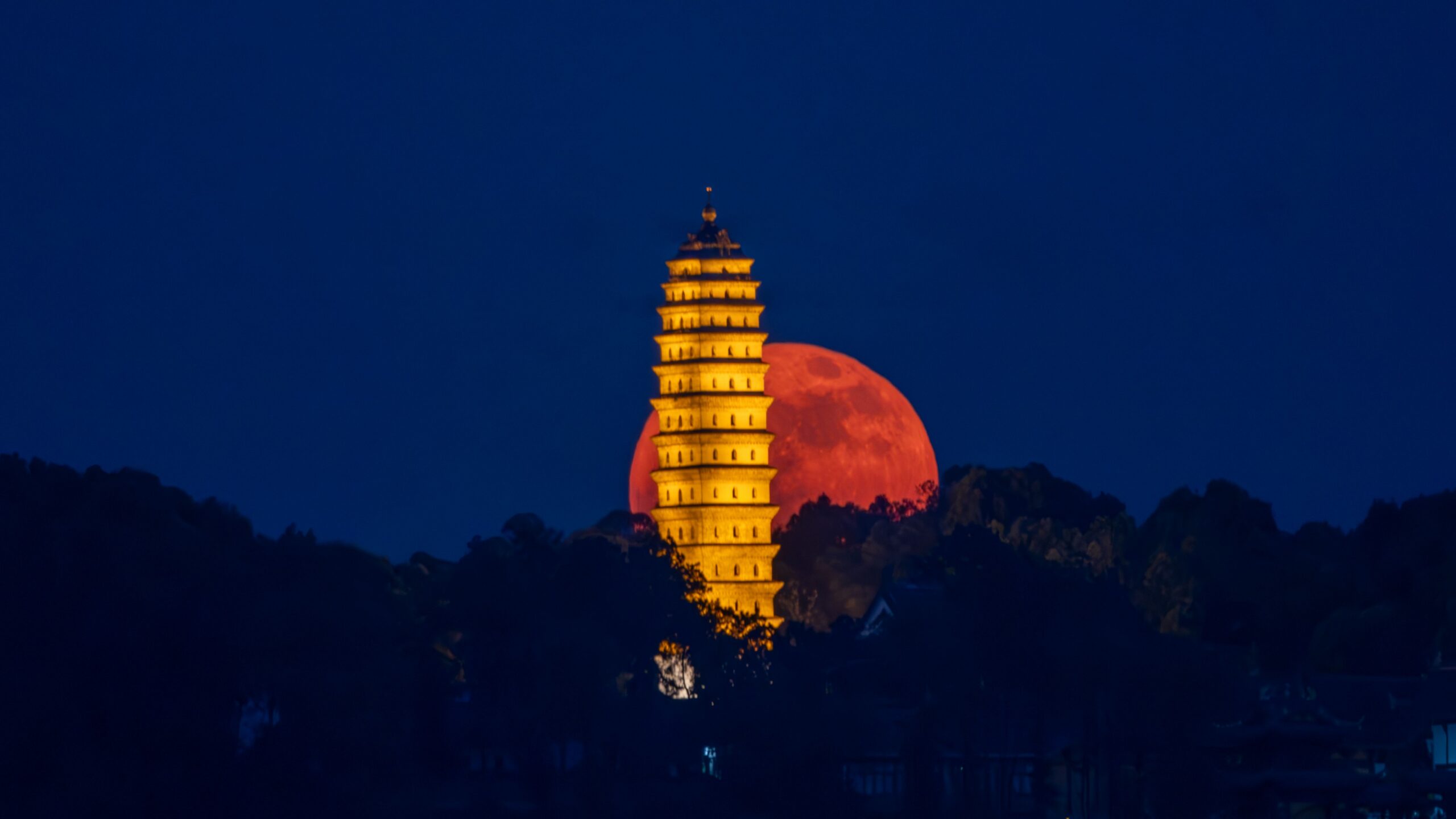 close up red strawberry moon low on the horizon behind a tall golden building.