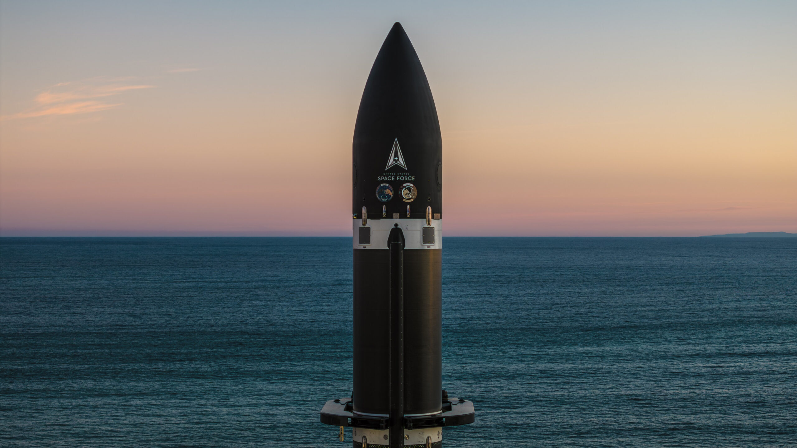 The top of a black rocket stands against a background of rich sea parted in the center by the horizon, and a cool, orange-hued sky.