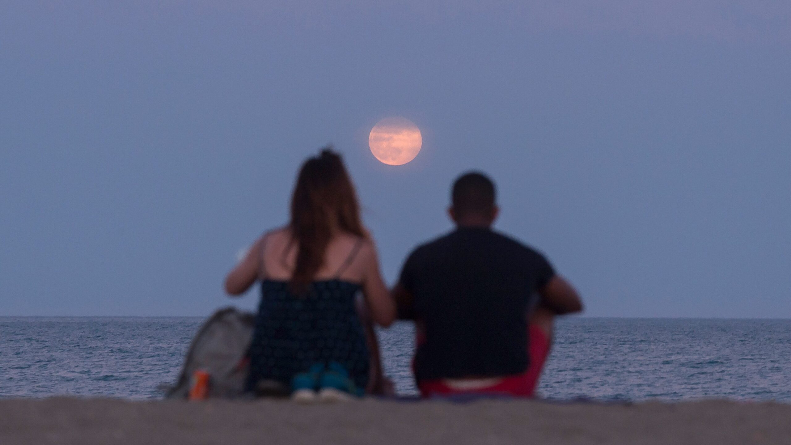 two people sit on a beach and look at a low hanging strawberry moon above the sea.