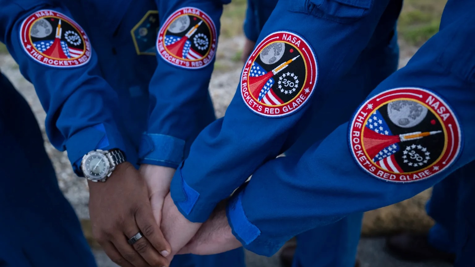 closeup photo of four blue-sleeved astronaut arms clasped together. on each sleeve is a circular, reddish mission patch
