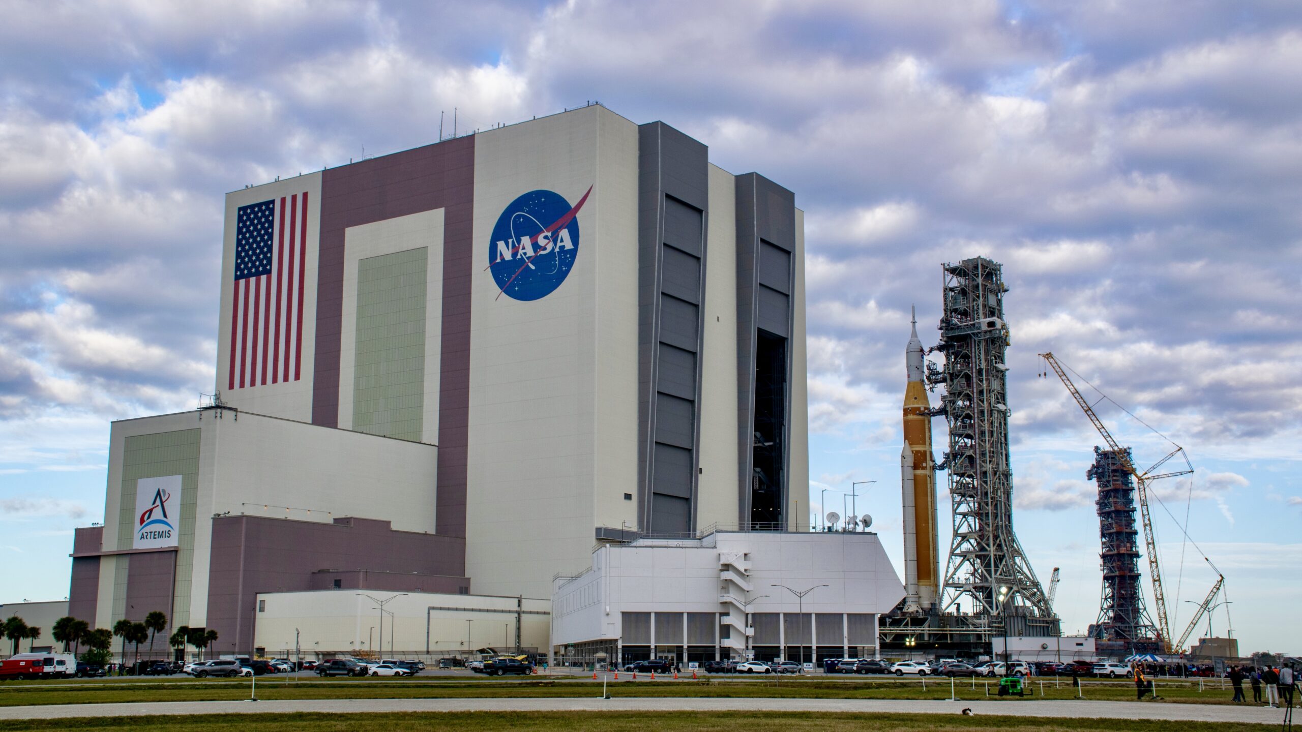 A bit orange rocket rolls out of a giant white building, with an extra launch tower in the background.