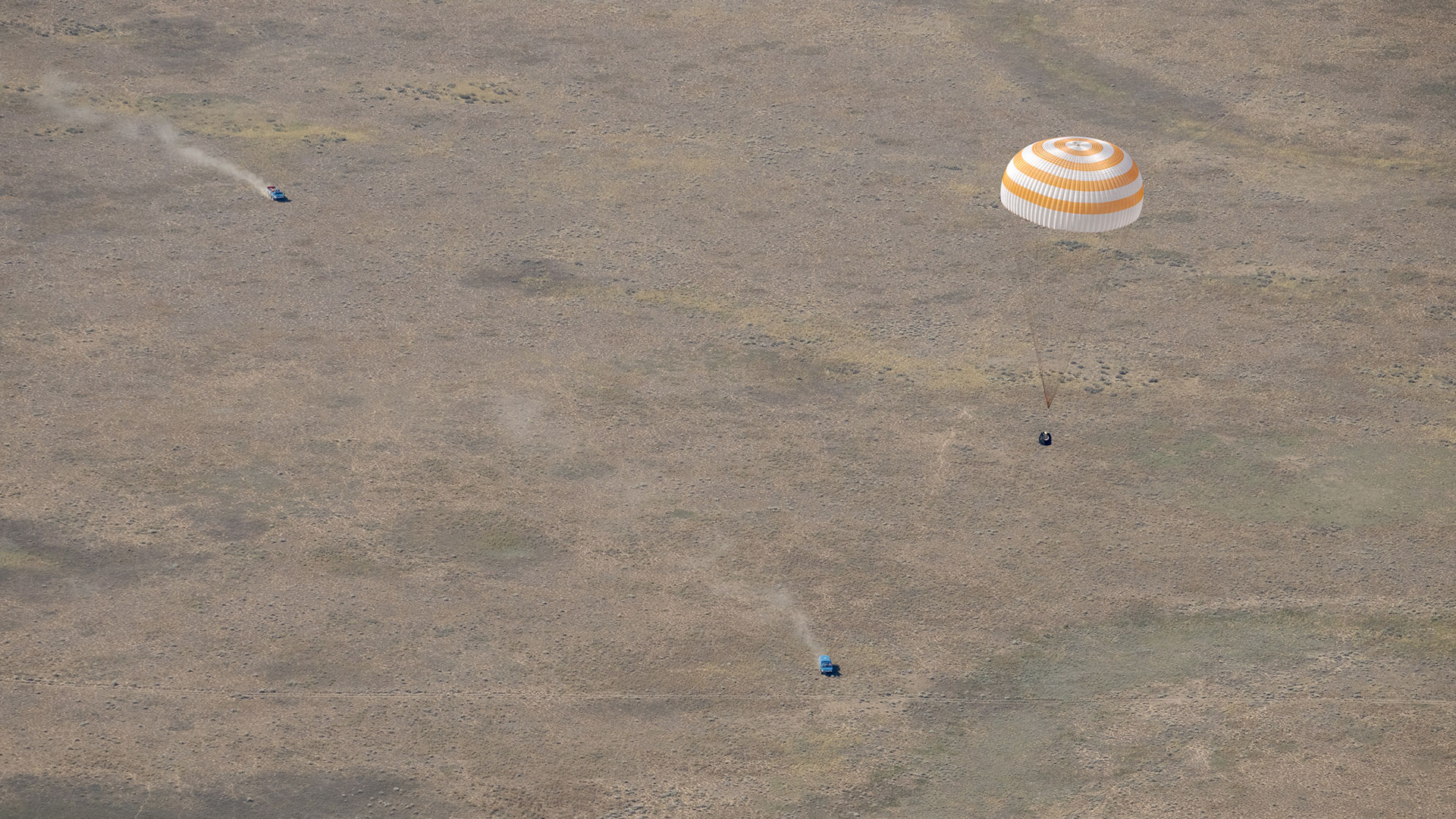 an aerial photograph of a space capsule descending under a large white and orange parachute to a landing on a barren steppe as recovery vehicles approach.