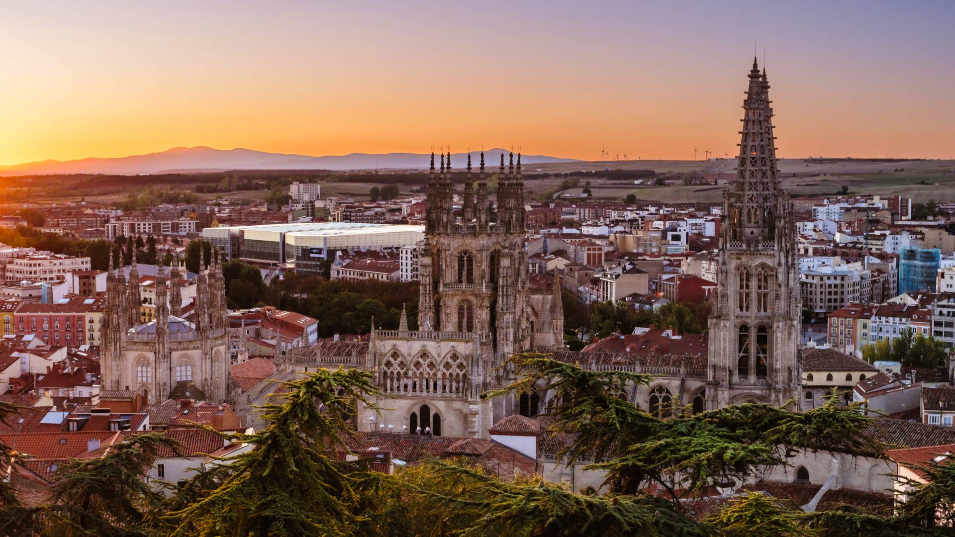 A panoramic view of a historic city at sunrise. There is a large Gothic cathedral with tall spires in the foreground and a mix of red-roofed buildings and greenery, with mountains in the distance.