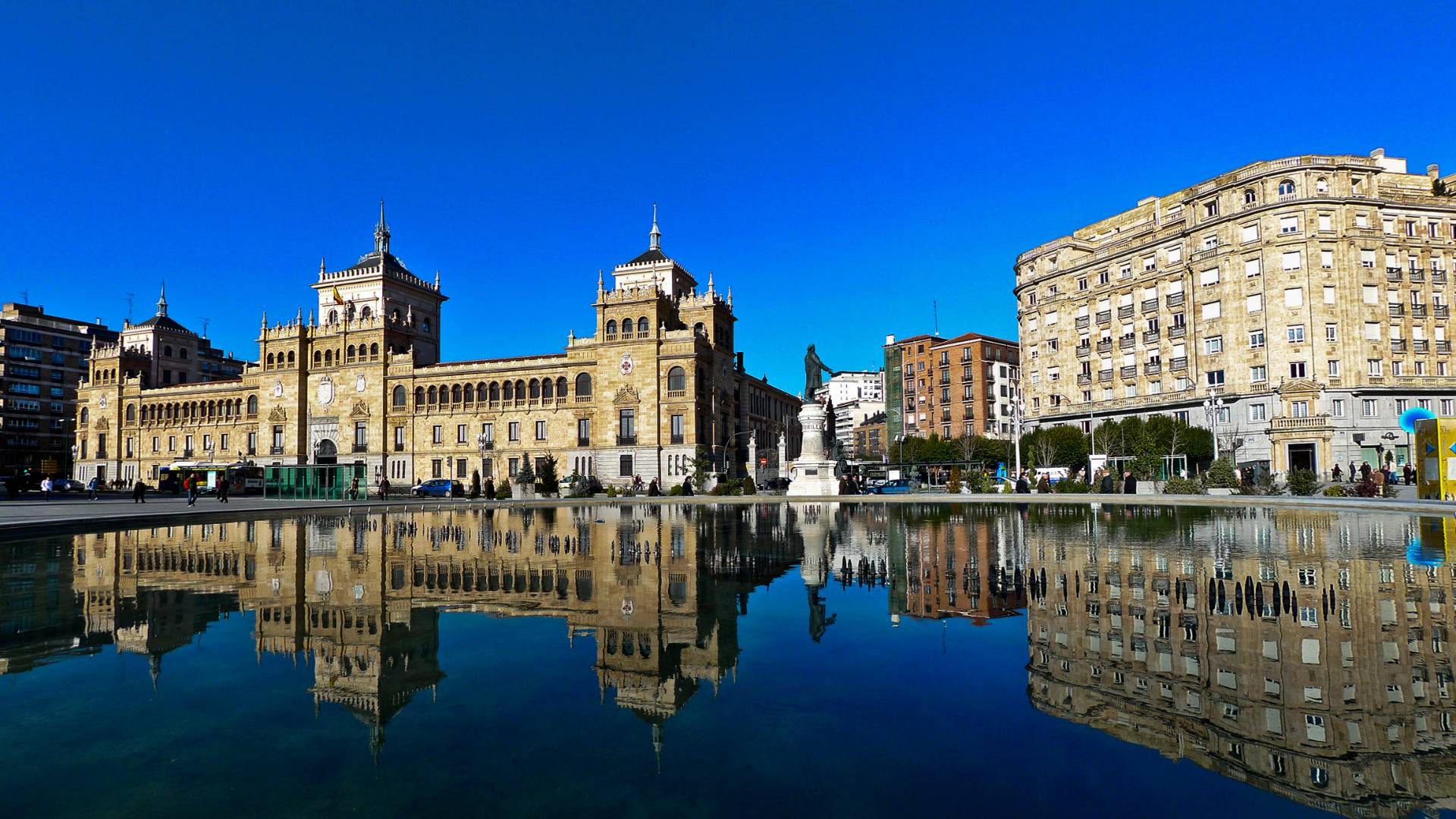 Large historic buildings are reflected in a big, still body of water in an urban plaza, all under a clear blue sky.