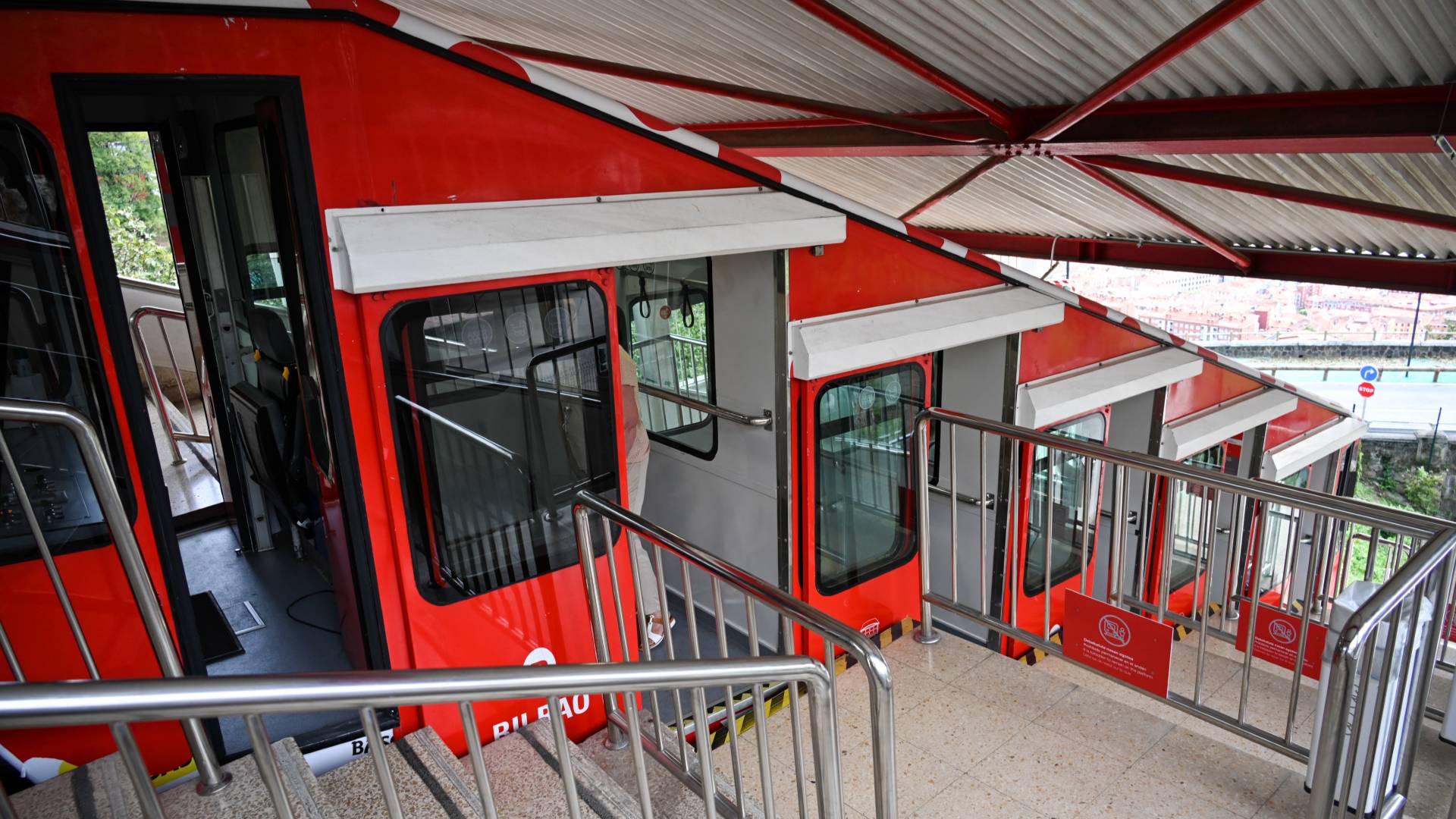 Red funicular train with multiple carriages parked at a station platform, with metal railings and a partially visible cityscape in the background.