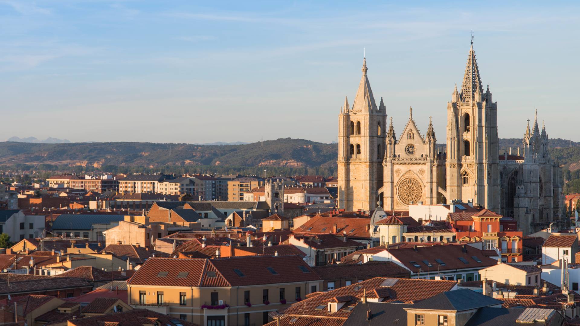 A panoramic view of a city with red-roofed buildings and the León Cathedral, a Gothic church with tall spires and ornate details, set against distant hills under a clear blue sky.
