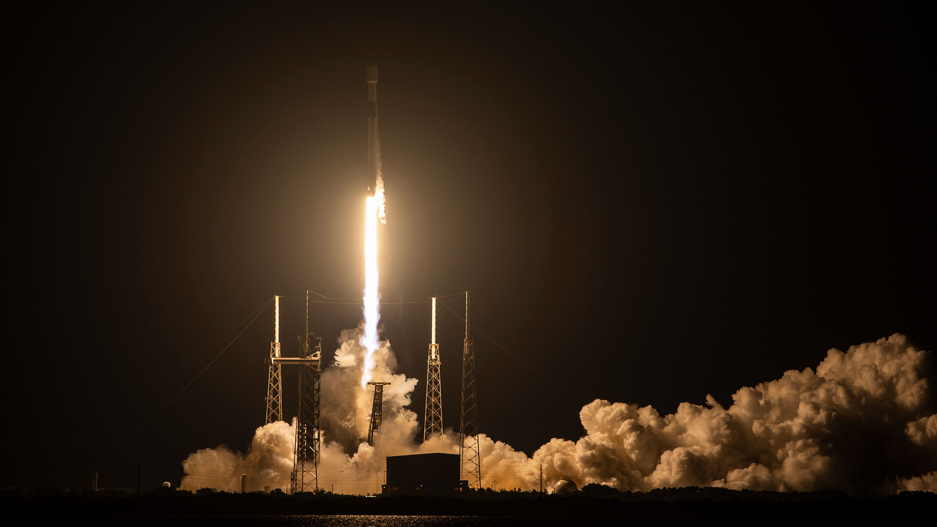 The bright orange-white plume from a rocket lights up its launch pad as it lifts off into the darkness of an early morning sky.
