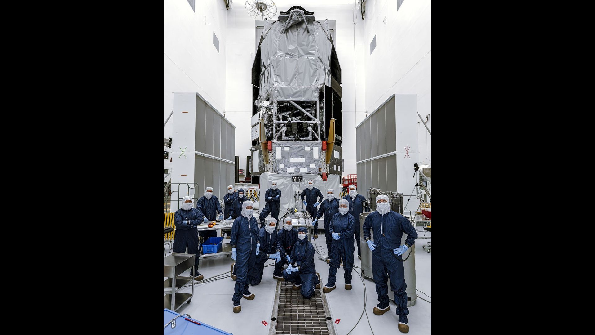 People in blue jumpsuits stand in front of a silver-wrapped telescope in a clean room.