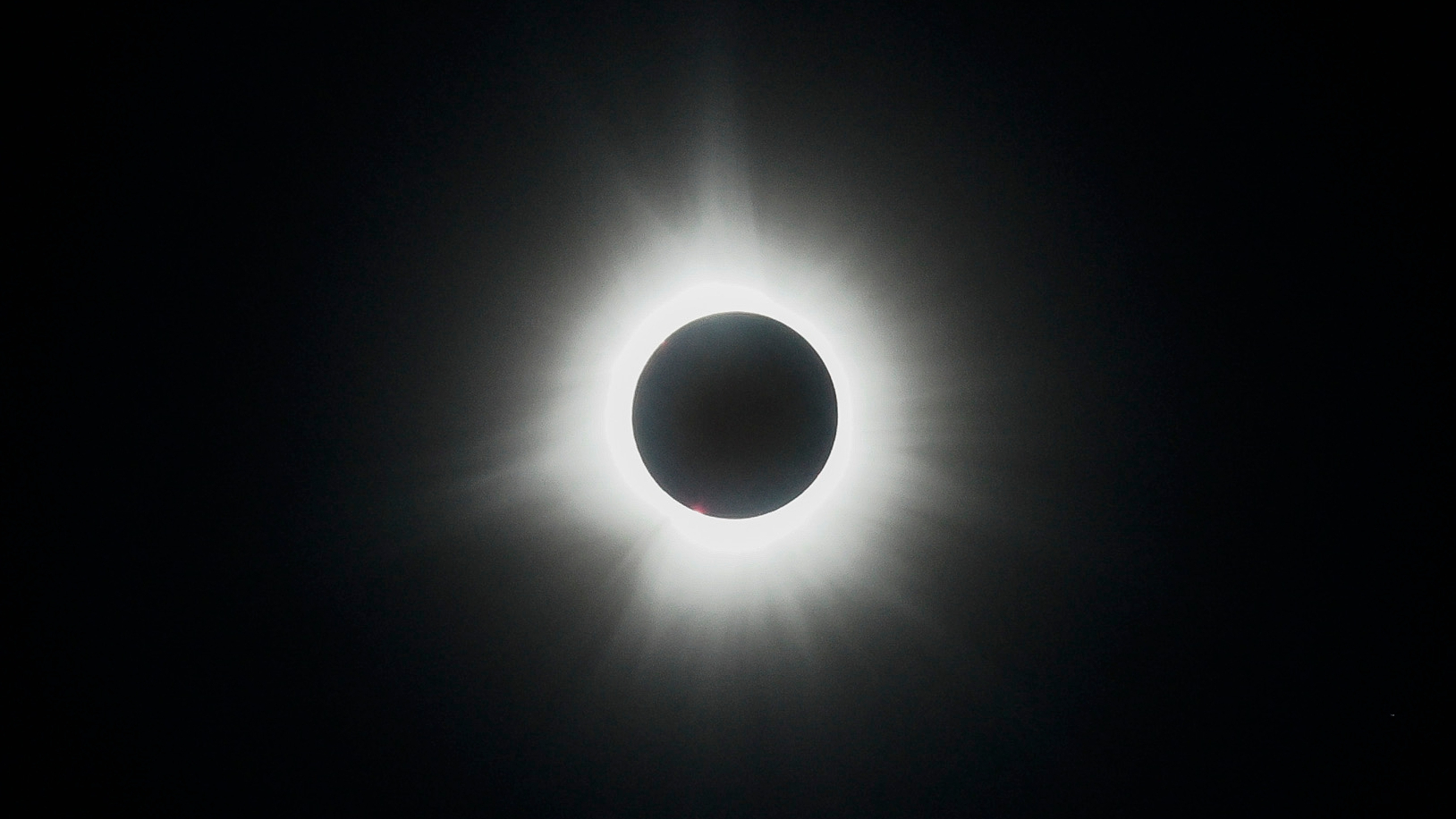 The moon's silhouette is visible in front of a fully eclipsed sun in a black sky, with the sun's white atmosphere creating a halo around the lunar disk.