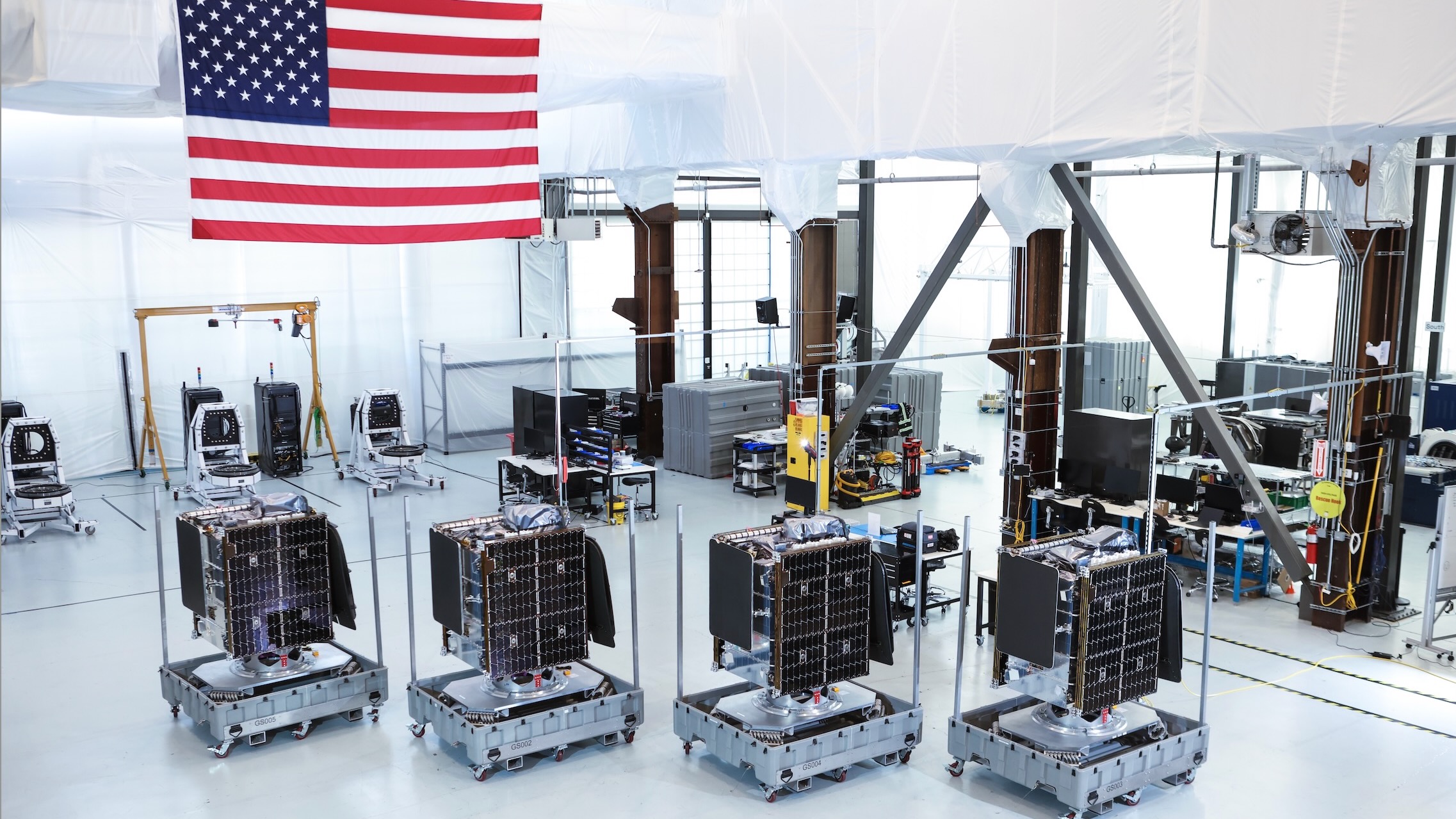 four cubical spacecraft sit inside a large white-walled clean room with an american flag in the background