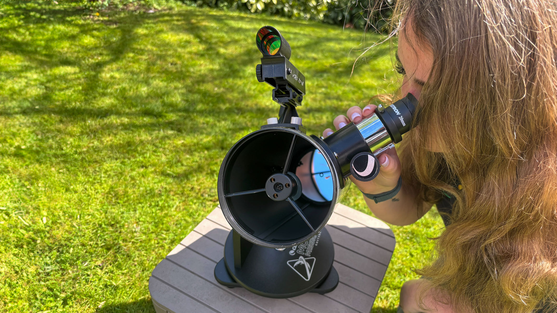 A woman looking through the Celestron Moon Mission 100mm Tabletop Dobsonian with grass in the background.