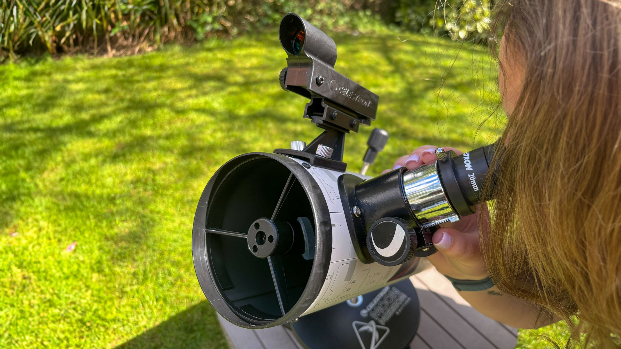 A woman looking through the eyepiece of the Celestron Moon Mission 100mm Tabletop Dobsonian with grass in the background.
