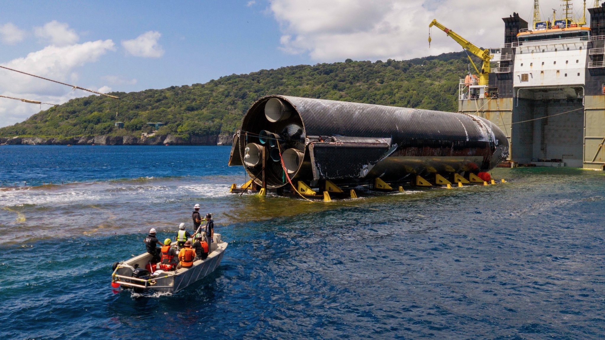 photo of a cylindrical gray spacecraft on the deck of a ship that's moored off a verdant island
