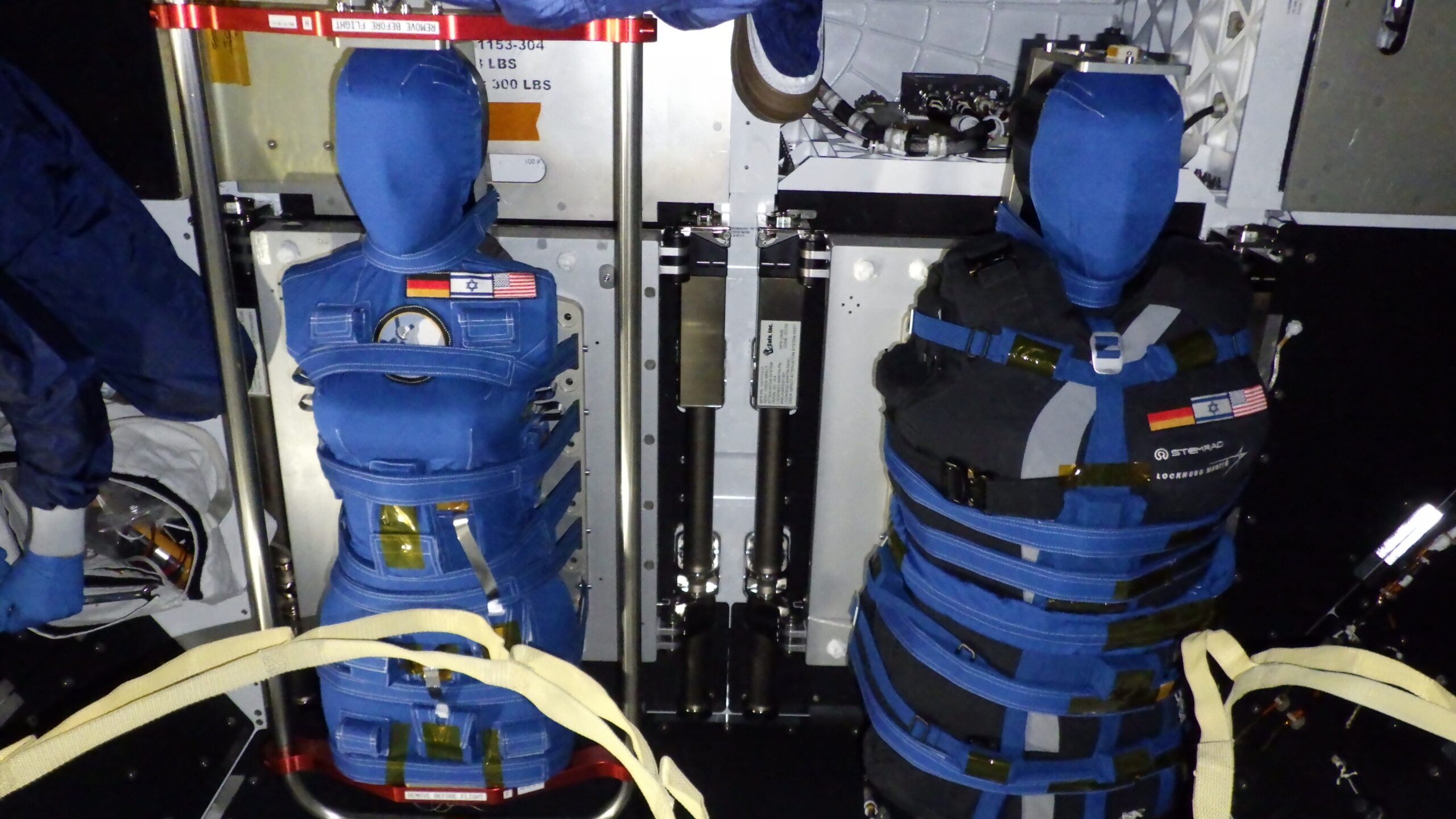 two blue-and-black torso mannequins stand upright inside a spacecraft, strapped in with blue bindings