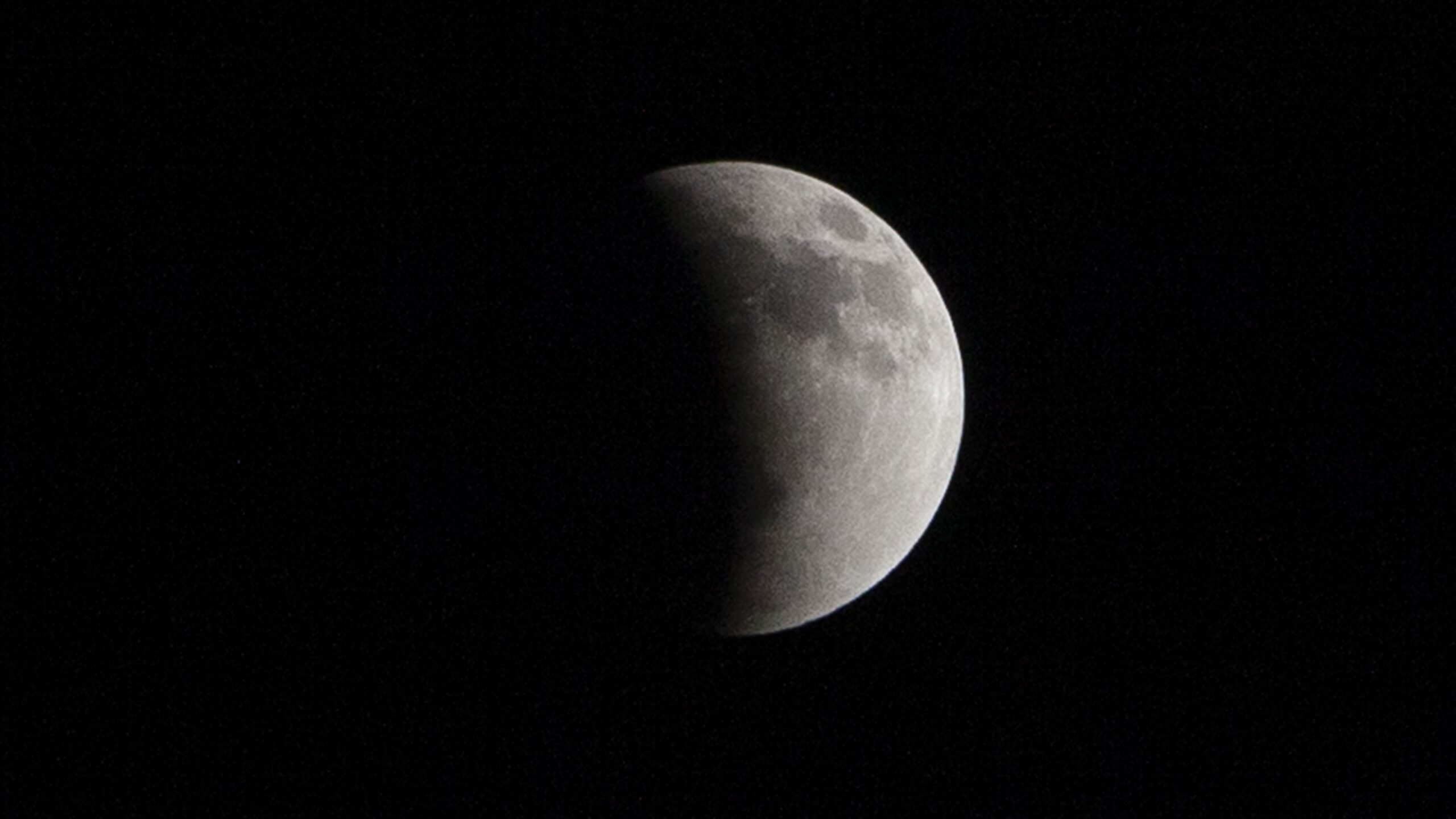 A full moon is photogrpahed in a black sky with its left half bathed in Earth's curved shadow during a lunar eclipse.