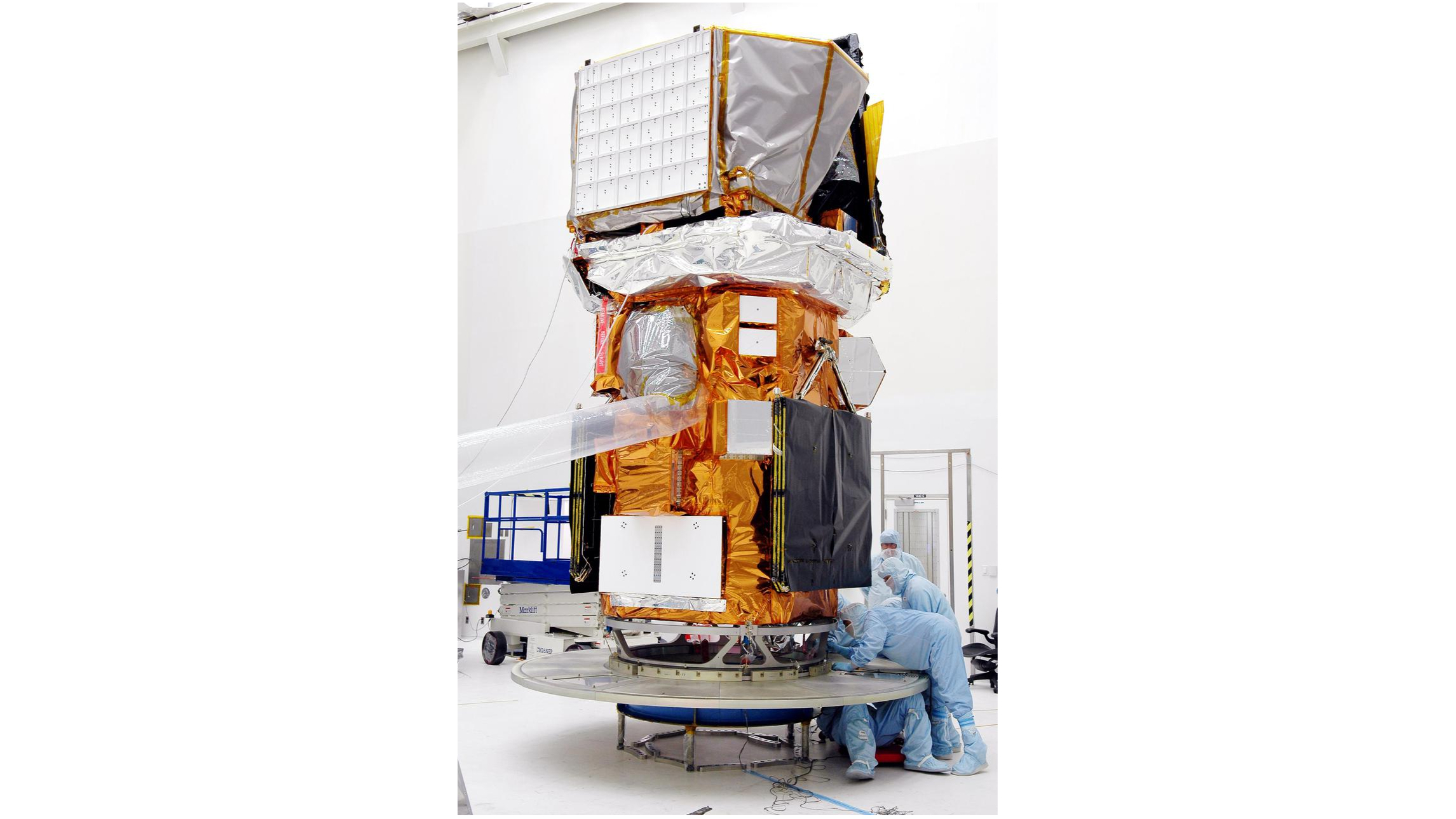 Three white-suited technicians work on a tall golden spacecraft in a big white clean room