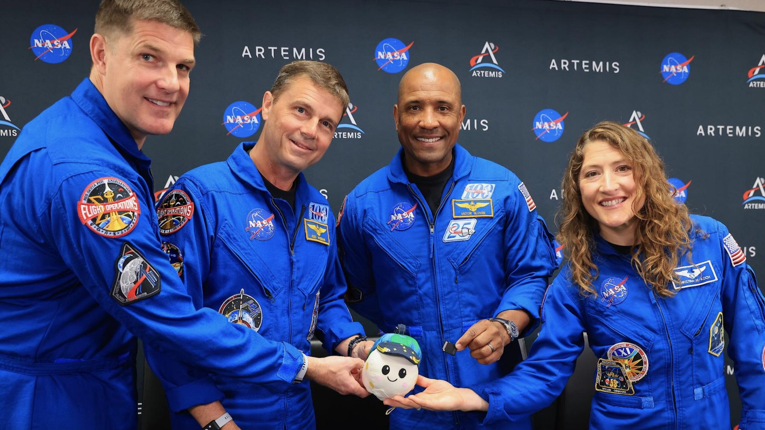 three male astronauts and a female astronaut, all wearing blue flight suits, stand in front of a gray wall