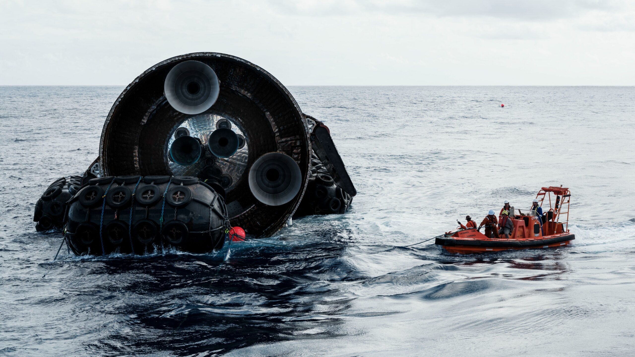 the aft end of a rocket with 3 small and 3 large engines floats in the ocean next to an orange raft with people aboard.