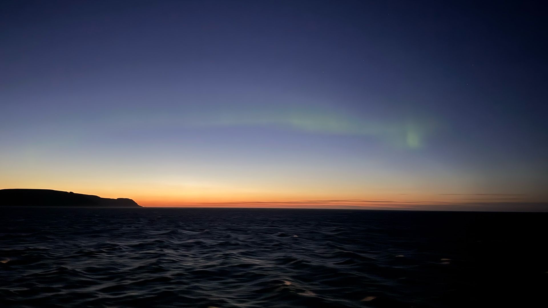 aurora visible in the sky as ribbon of green light during twilight hours with the glow of the sunset on the horizon.