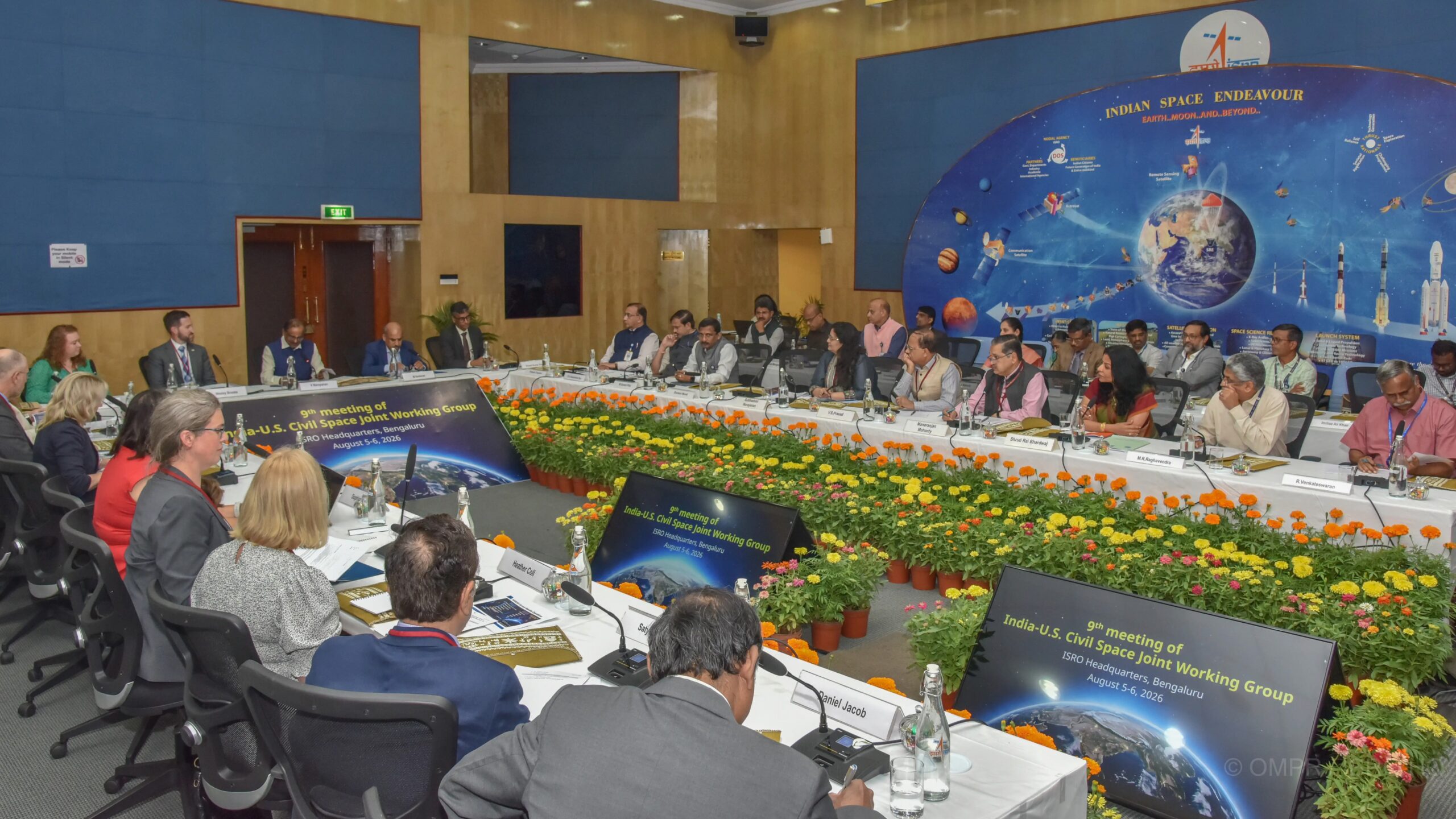 about 30 well-dressed people sit around a huge u-shaped table decorated with flowers, during a space conference