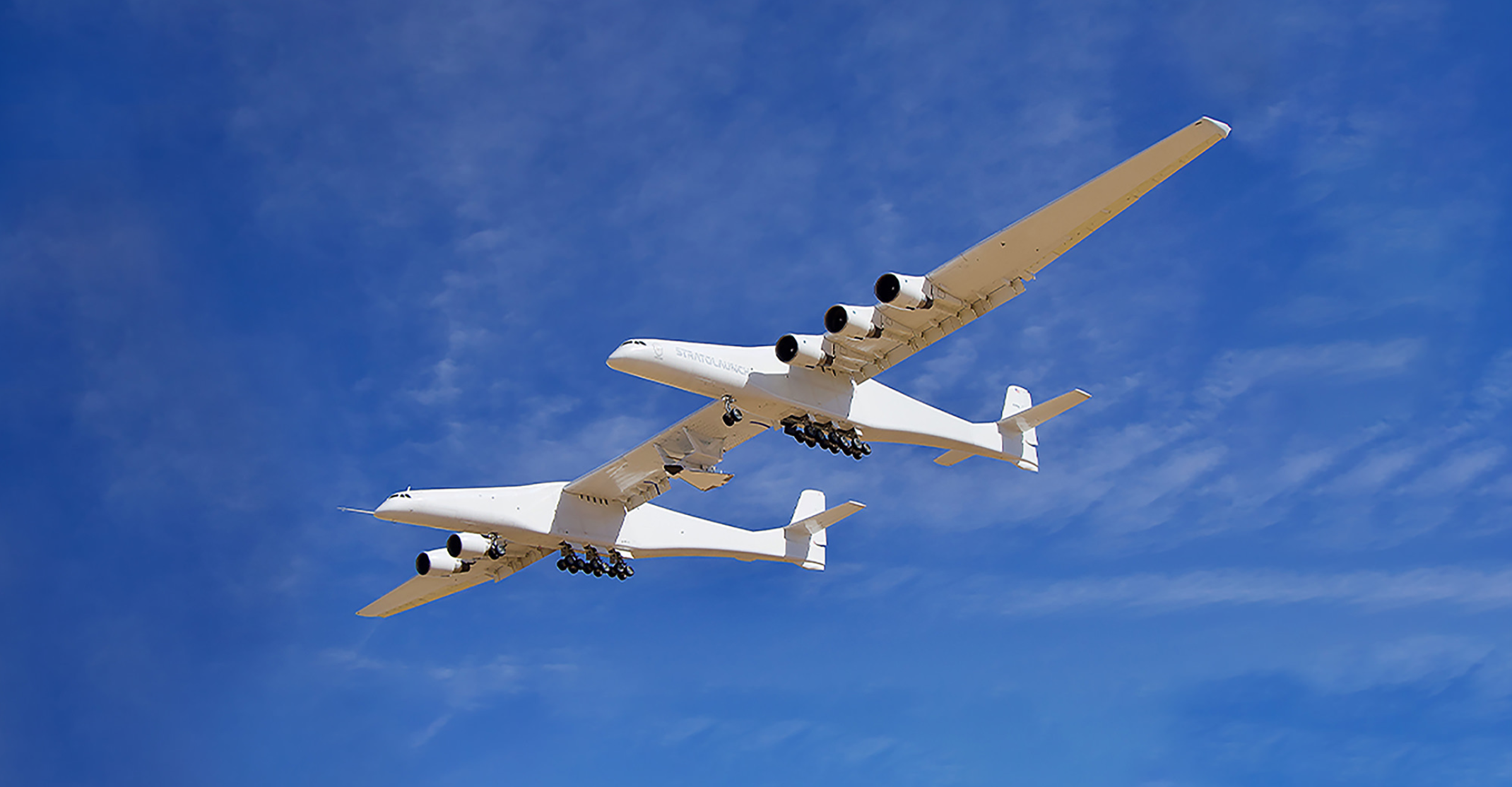 a white dual-fuselage aircraft with an experimental rocket plane under its wing