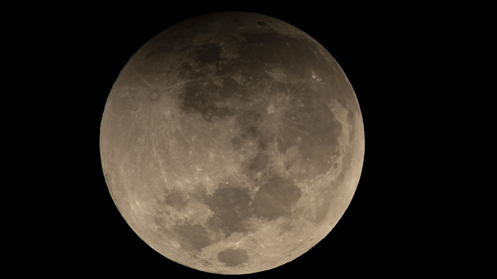 A full moon is shown in a black sky with its upper edge shadowed during a partial lunar eclipse.