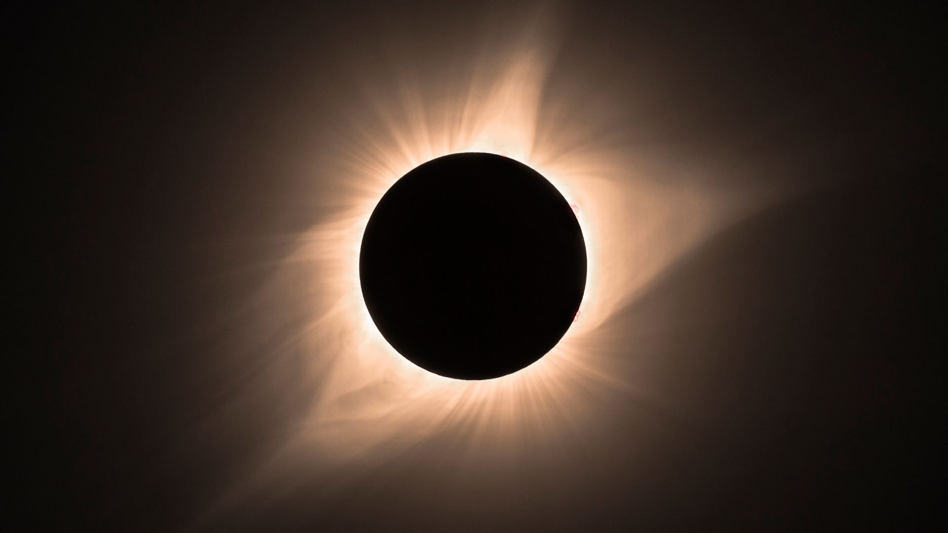 A fully eclipsed sun is photographed at the height of a total eclipse in a black sky, with its golden atmosphere streaming away from the lunar disk.