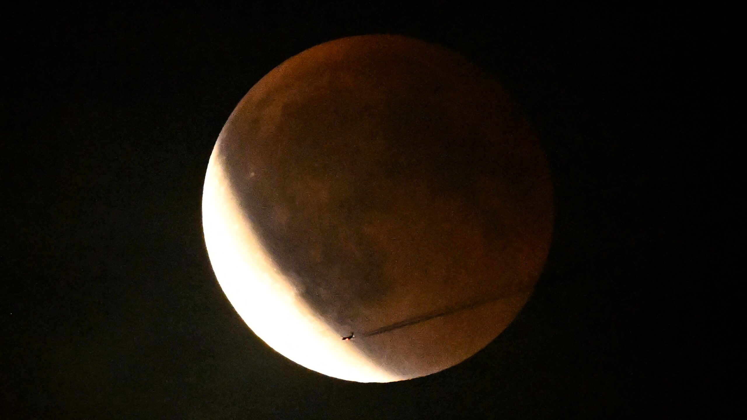 A full moon is shown during a lunar eclipse as Earth's shadow covers its right side, turning it a subtle red, as a silver arc shines to the lower right.