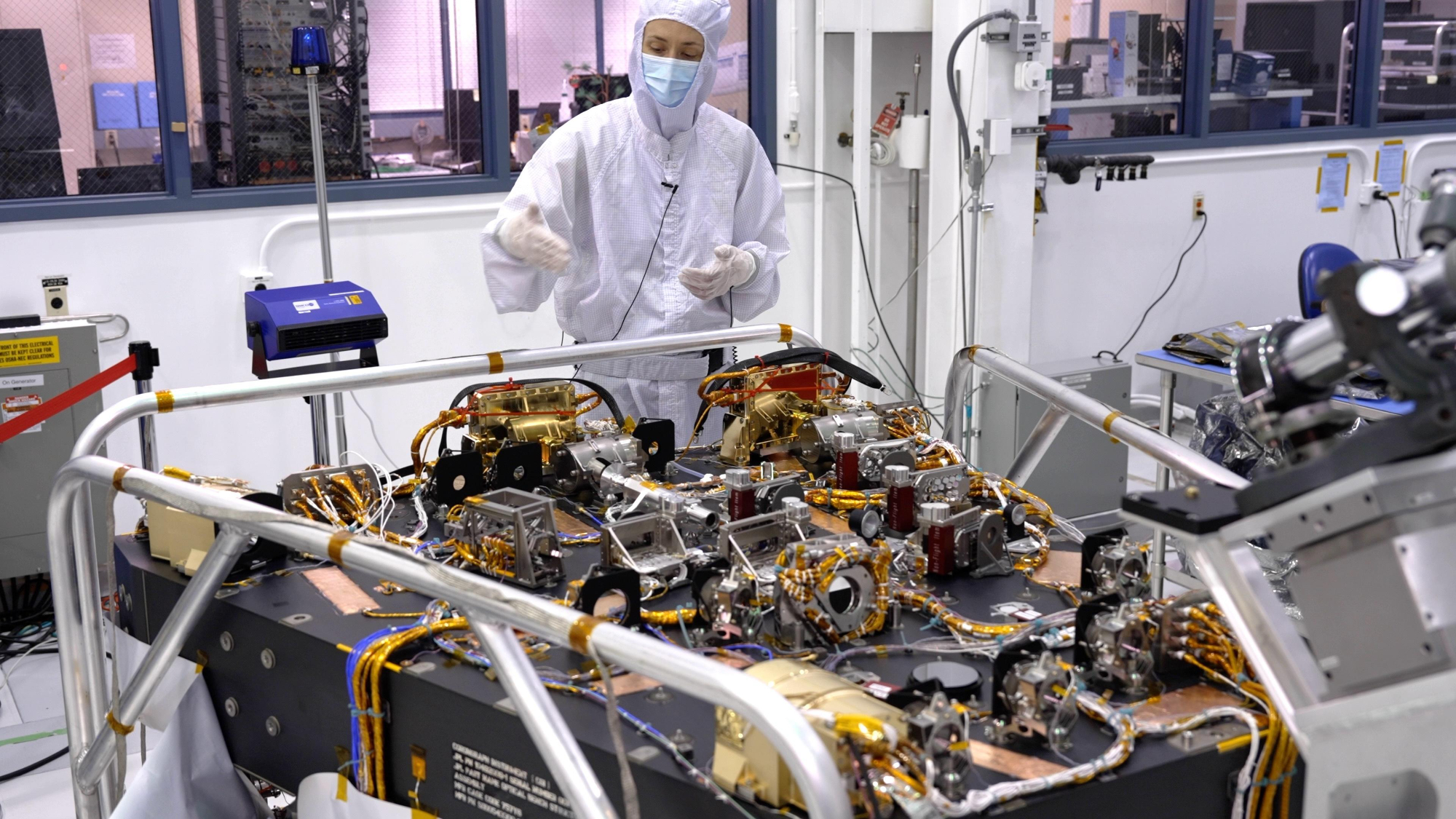A bunch of golden components and wires in top of a large black box. A person in a white clean room suit stands behind the contraption.