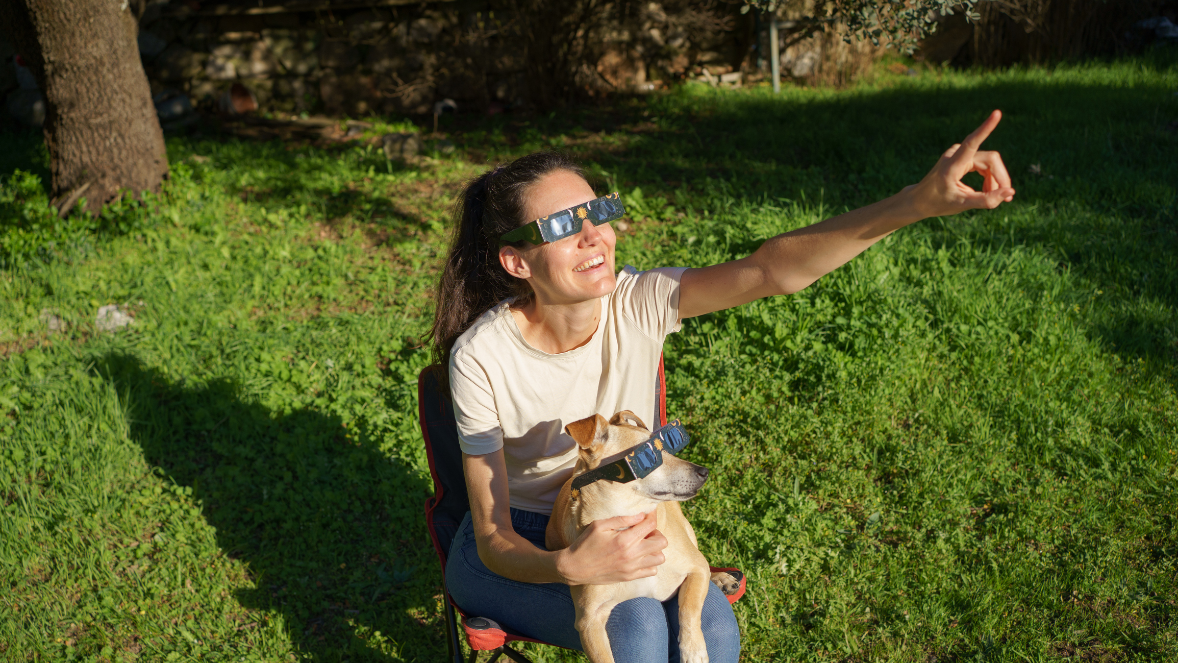a woman wearing solar eclipse glasses smiles and points up to the sky. There is a dog on her lap also wearing solar eclipse glasses and looking towards the sky.