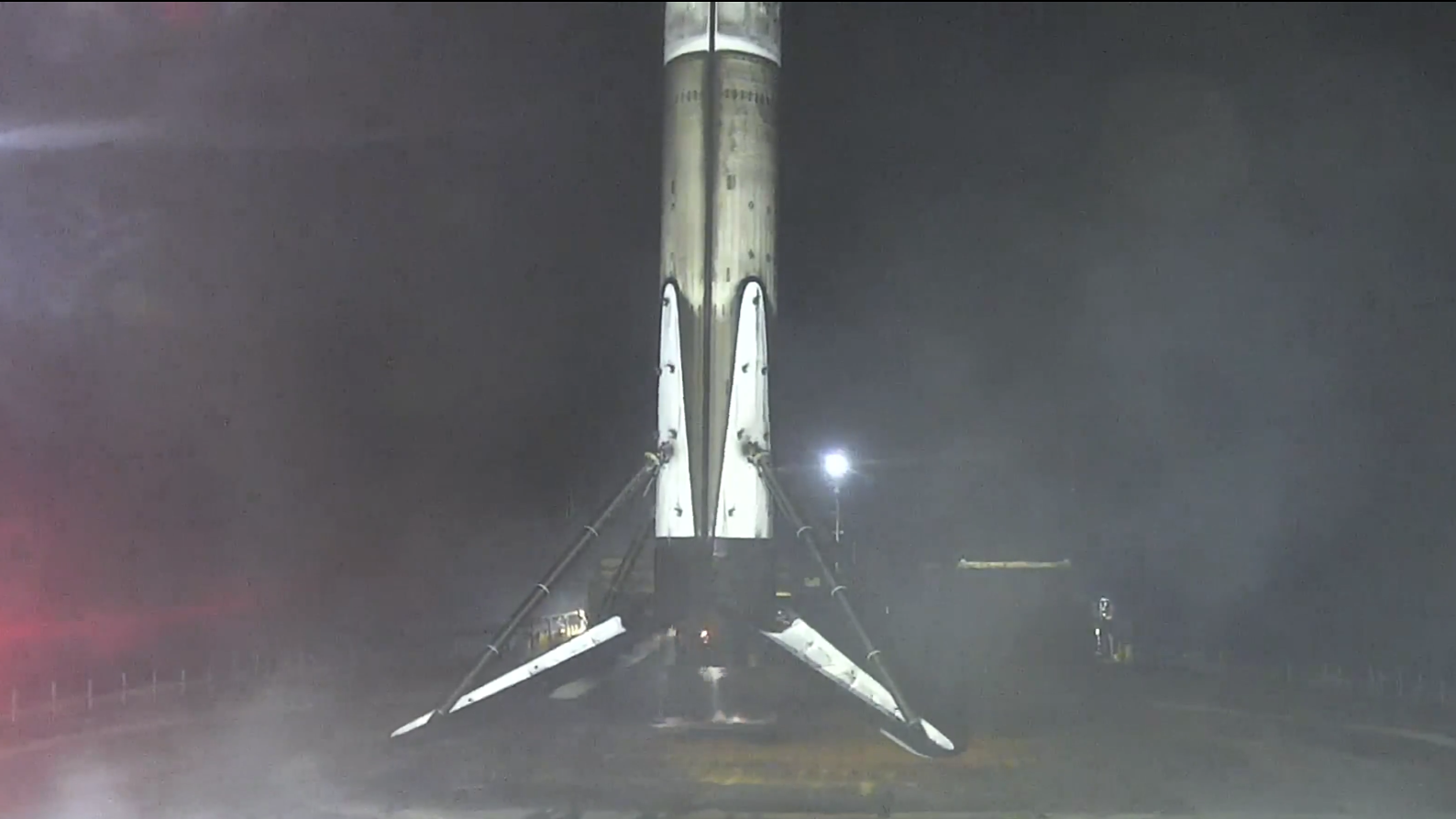 A charred white rocket stands with legs extended on a dark barge at sea.