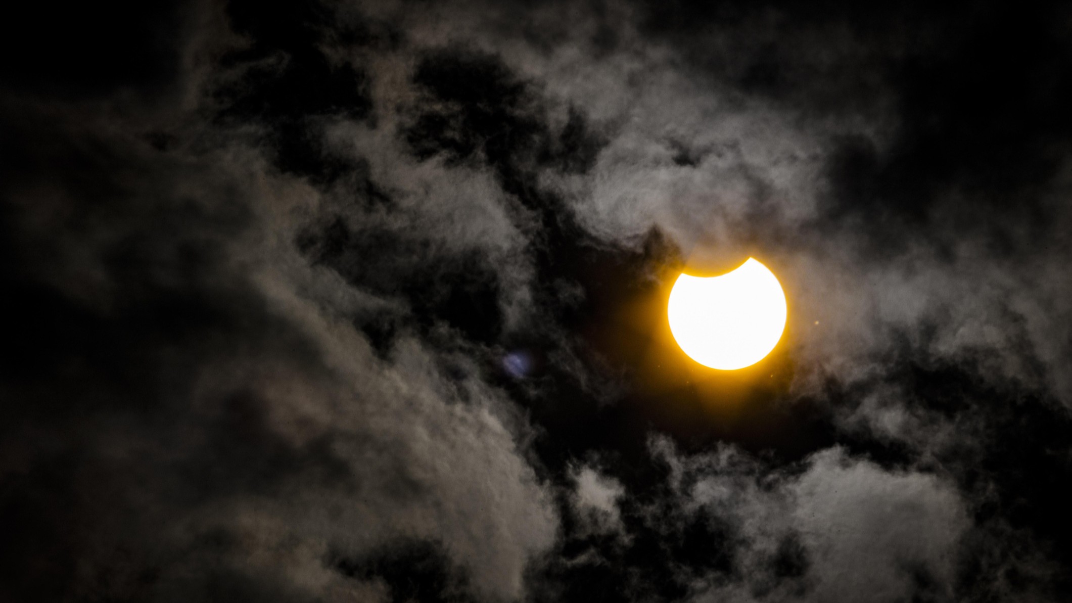partial solar eclipse shows the upper portion of the sun covered by the moon, surrounded by partly cloudy skies.