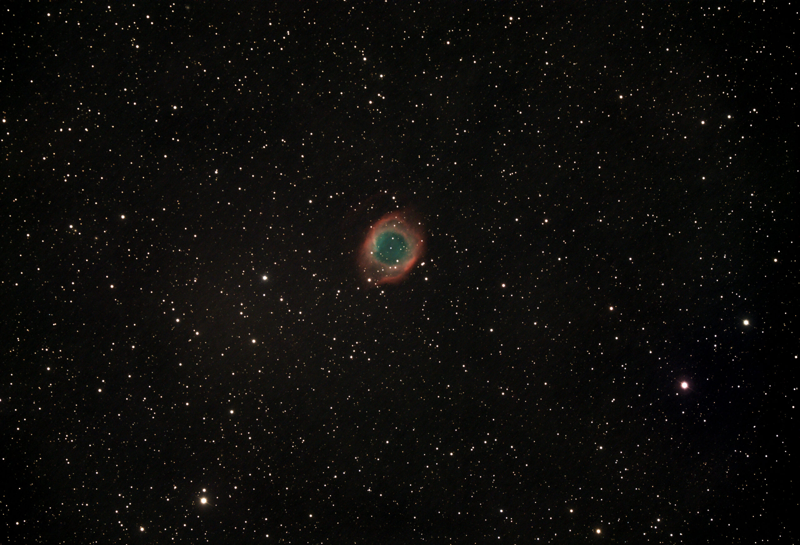 A telescopic photograph of the Helix Nebula (NGC 7293), a large planetary nebula appearing as a circular ring of glowing teal-green gas surrounded by a diffuse red outer halo, set against a black sky scattered with orange and white foreground stars.