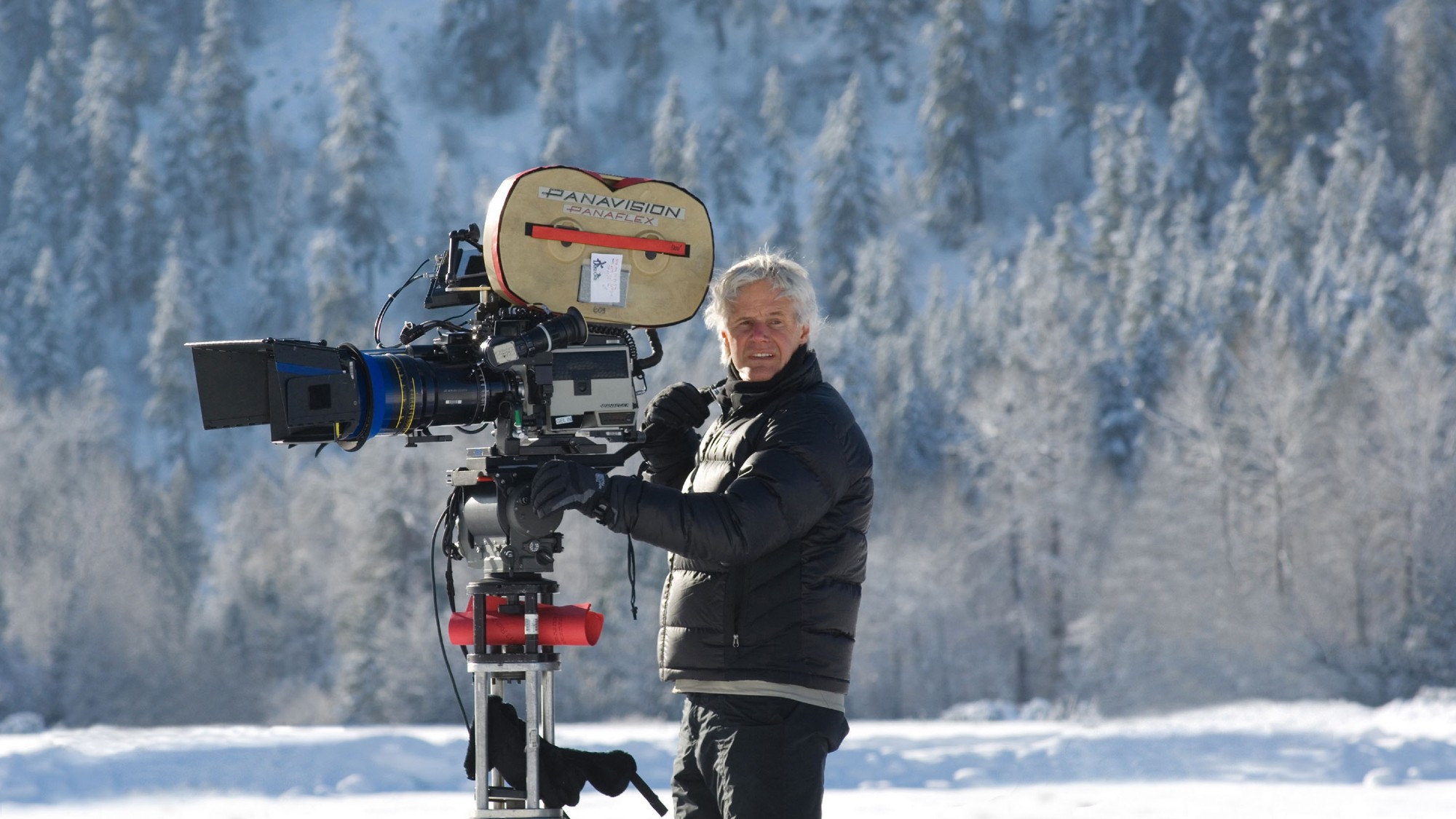 a man in winter clothing holding a large film camera outside in a snowy forest landscape