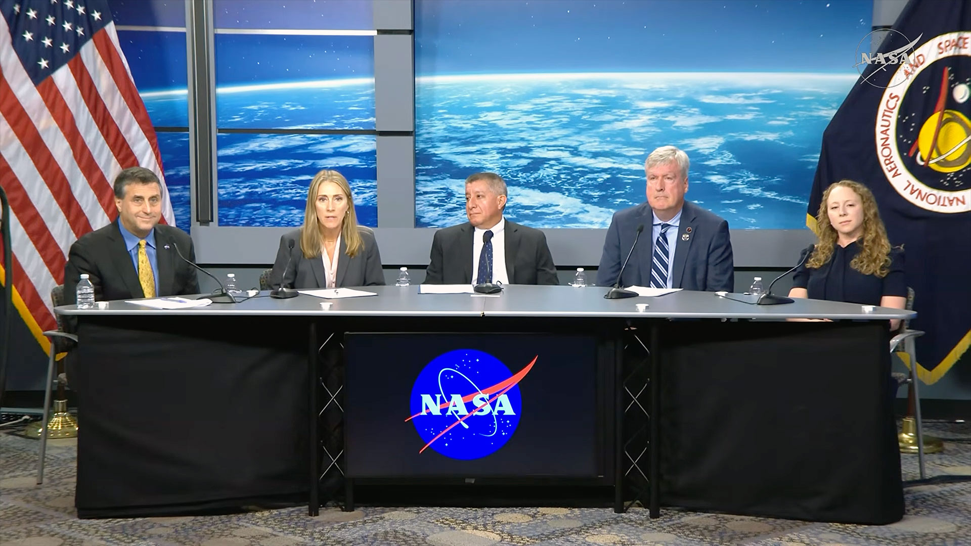 two women and three men sit wearing business attire sit at a table with a NASA logo during a press conference