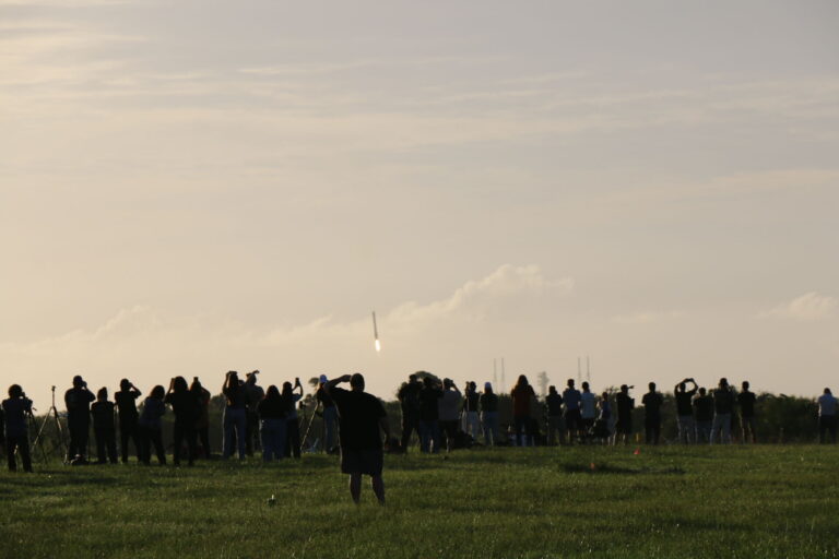 People stand on a lawn watching a long structure descend to Earth.
