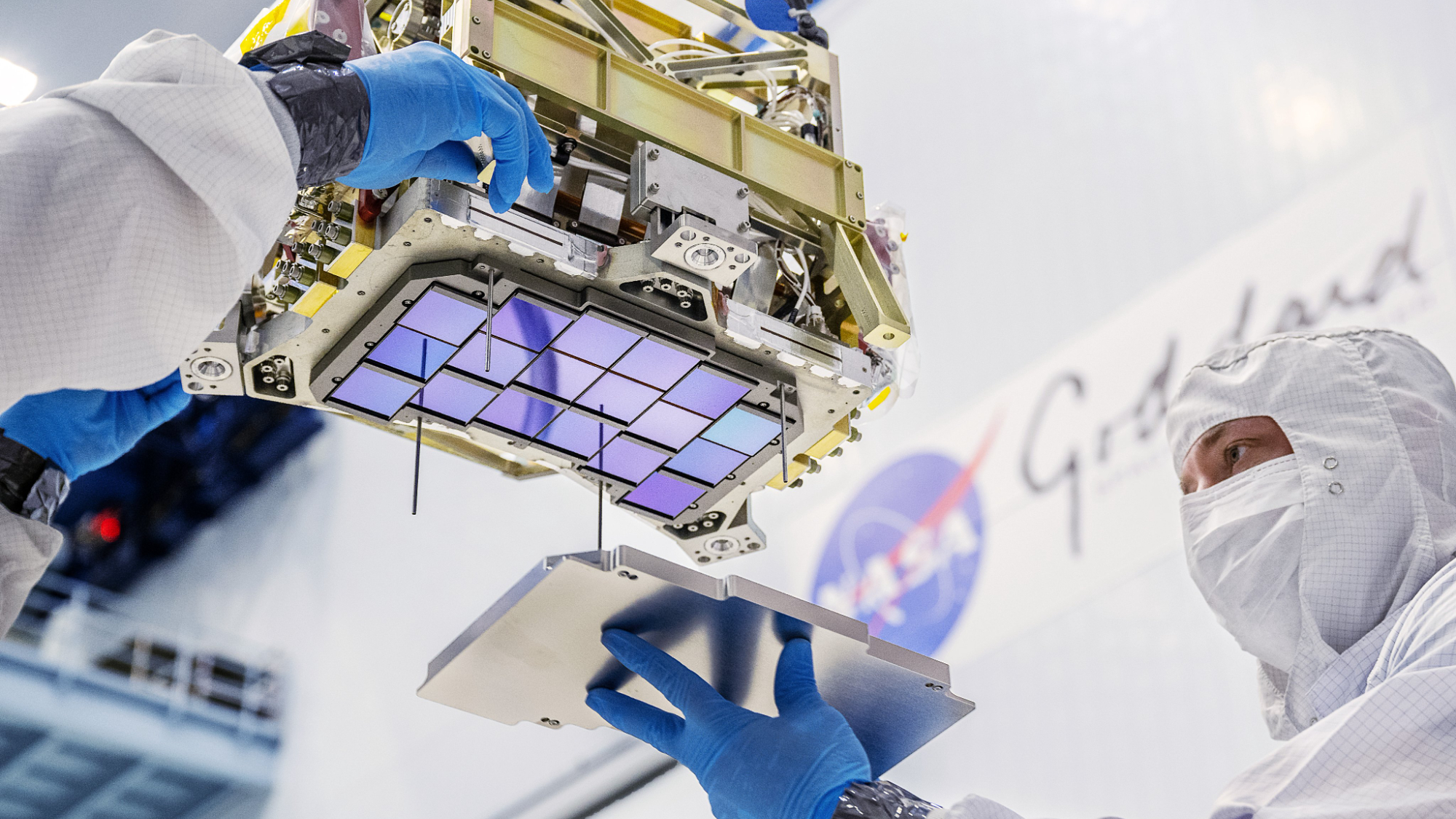 Blue reflective squares are attached to an instrument that hangs from above. A person in a white clean room suit is putting a metal door on top of the reflective squares.