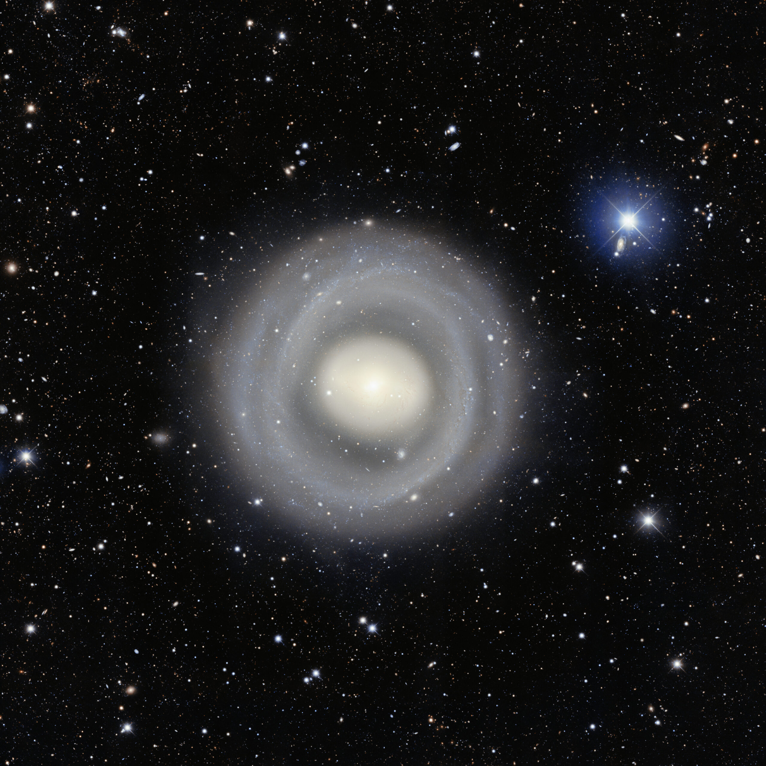 A telescopic photograph of a nearly face-on ringed spiral galaxy with a bright, glowing white-yellow core surrounded by multiple faint concentric blue-white spiral rings extending outward. The field is rich with distant background galaxies of various shapes and sizes scattered across the black sky, and a bright foreground star with prominent blue diffraction spikes is visible in the upper right.