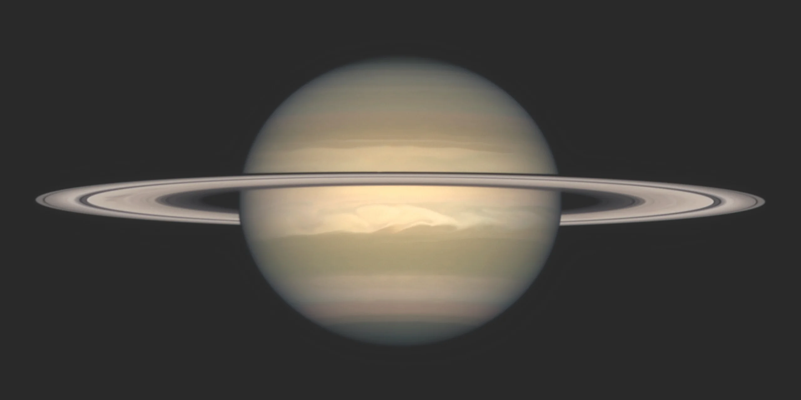 Photograph of Saturn against a dark background, seen nearly edge-on with its ring system stretching horizontally across the globe and casting a thin shadow band on the planet. The pale gold and tan cloud bands are visible, with a bright white storm feature swirling in the planet's southern hemisphere.