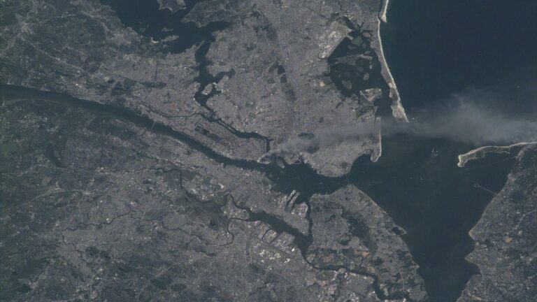 satellite photo of a large city on a bay, with a river running through it and smoke rising from the center of the city