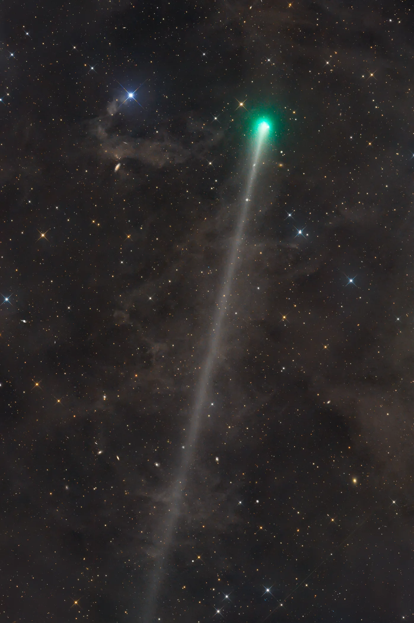 A bright green comet head glows near the top of a star-filled frame, streaming a long, faint white tail that stretches diagonally down almost the full height of the image. Wisps of dim brown dust clouds drift across the dark background, with a vivid blue-white star near the upper left.