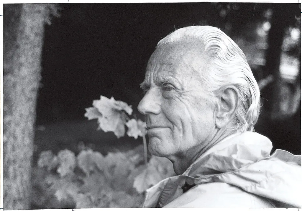 A black-and-white profile portrait shows an elderly man with slicked-back white hair looking toward the left side of the frame. Bright daylight highlights his face and light-colored jacket collar, contrasting against a dark, shaded background featuring a tree trunk and out-of-focus foliage.