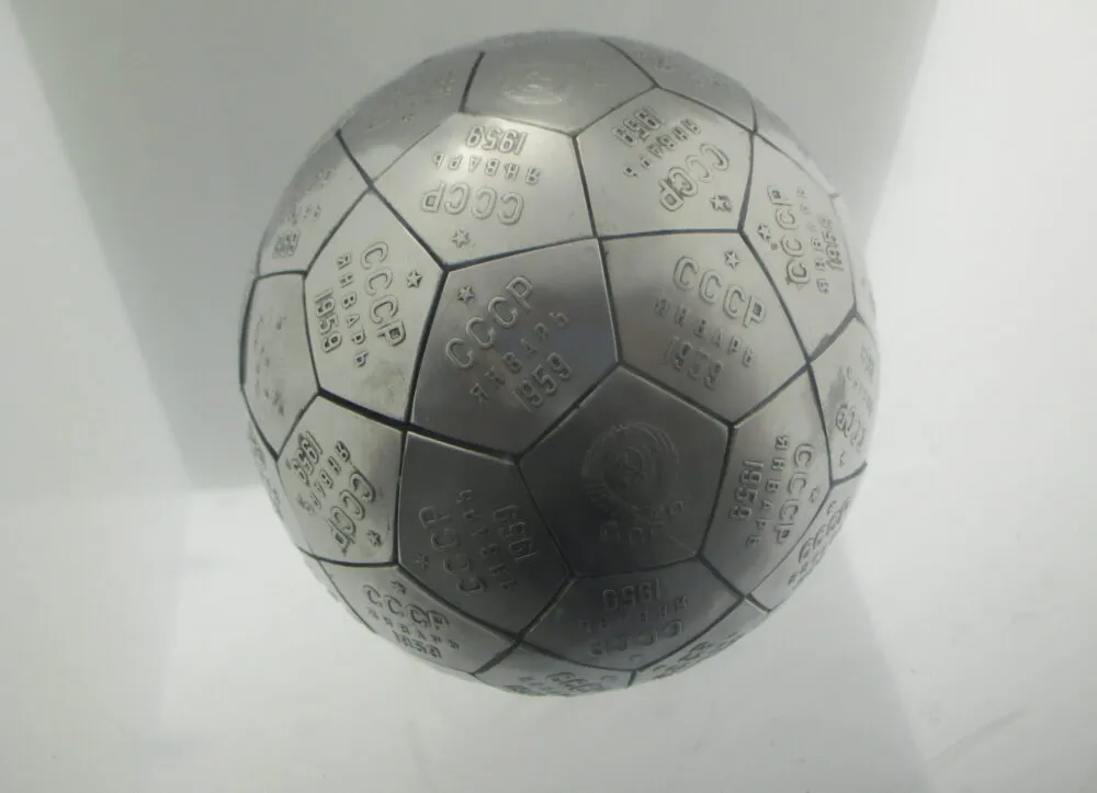 A metallic grey sphere made of interlocking pentagonal and hexagonal tiles sits centrally against a bright, plain white background. Each brushed-metal tile is engraved with Cyrillic text, numerals, and small crest emblems, resembling a soccer ball structure. Overhead display lighting creates soft reflections on the upper hemisphere and subtle shadows along the lower curves.